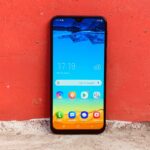 Galaxy M20 em pé com a tela ligada, encostado em uma parede vermelha.