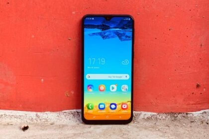 Galaxy M20 em pé com a tela ligada, encostado em uma parede vermelha.